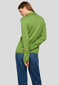 Grüner Strickpullover mit rundem Ausschnitt und langen Ärmeln, kombiniert mit dunkelblauen Jeans. Glatte Textur und figurbetontes Design.