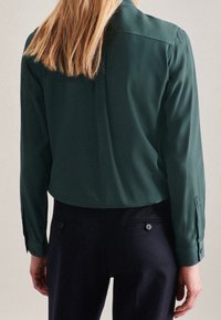Donkergroene lange mouwen shirt met een gladde textuur, rug naad, en knoopmanchetten, gecombineerd met donkere broeken met achterzakken.