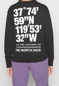 Černá mikina s velkým bílým textem na zadní straně, kter�á obsahuje geografické souřadnice a značení "THE NORTH FACE". Měkká tkanina a volný střih.