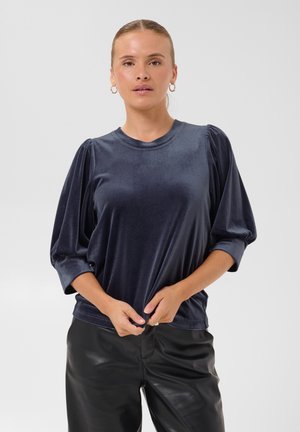 Le mannequin porte un haut en velours bleu marine foncé à manches bouffantes et un pantalon noir, et tient l’ourlet devant un fond clair uni.