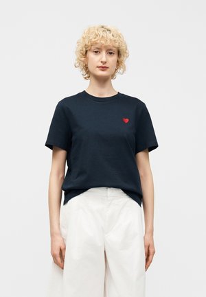 Personne blonde portant une chemise noire à manches courtes avec un petit emblème en forme de cœur rouge sur la poitrine et un pantalon blanc taille haute.