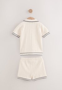 Strikket todelt sæt i creme, med en kortærmet polo-top med marineblå detaljer og matchende shorts, begge med ribstrikket tekstur.