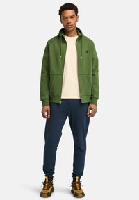 Grüne Zip-Hoodie mit Kordelzügen, getragen über einem beigen T-Shirt. Kombiniert mit navy Jogginghosen und braunen Schuhen mit gelben Akzenten.