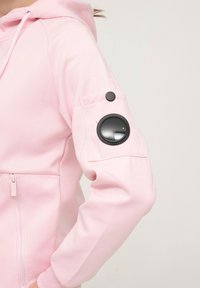 Sweatshirt rose avec une fermeture éclair sur le devant, une poche kangourou et un accent noir rond sur la manche avec un détail de bouton. Texture douce.