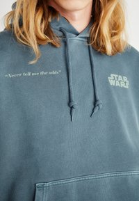 Hoodies pasztell kékkal, osztott kenguru zsebbel. A "Soha ne mondd el nekem az esélyeket" és a "STAR WARS" grafika kontrasztos színekben.