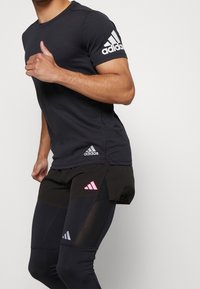 Svart atletisk T-shirt med korta ärmar, med en vit Adidas-logga på ärmen och en åtsittande design för aktiv träning.