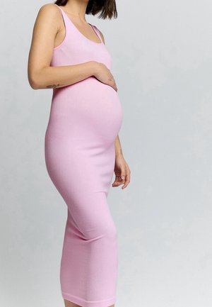 Une femme enceinte, vêtue d’une robe rose ajustée sans manches, pose une main sur son ventre devant un fond gris uni.