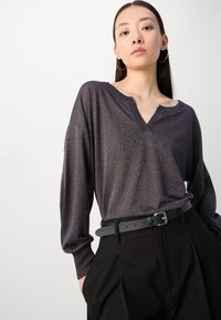 Langärmlige V-Ausschnitt-Bluse aus dunkel metallisch lila Stoff, mit lockerem Schnitt und geripptem Ausschnitt, kombiniert mit hochgeschnittenen schwarzen Hosen.