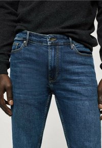 Tummansininen farkkuhousut kapealla istuvuudella, joissa on etukappaleen nappikiinnitys, viisi taskua ja hillityt ompelu-yksityiskohdat.