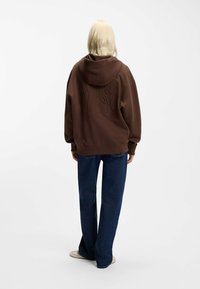 Bruine hoodie met capuchon en een textuurverhoogd ontwerp aan de achterkant. Gecombineerd met blauwe brede jeans en beige schoenen. Eenvoudige, ontspannen pasvorm.