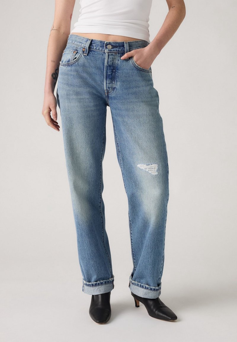 Personne portant un jean bleu clair retroussé avec une pièce usée sur la cuisse droite, un débardeur court blanc, et des bottes noires à talons.