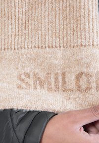 Beige strikket tørklæde med ribbet tekstur, prydet med ordet "SMUL" i en lidt mørkere nuance. Blødt og varmt materiale, holdt af en hånd.
