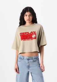 Afkortet beige t-shirt med vid hals og korte ærmer. Har fed rød tekst og logo-tryk centreret foran. Bæres med blå jeans.
