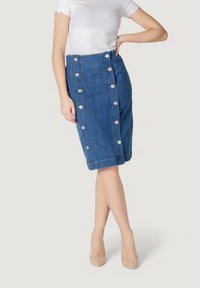 Falda lápiz de mezclilla en azul medio con detalle de botones laterales, a la altura de la rodilla y con un ligero desgarro en el dobladillo. Combinada con una blusa ajustada blanca.