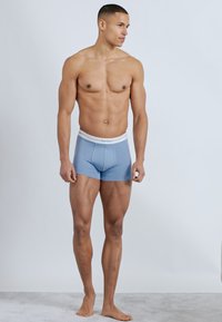 Άνδρας μοντέλο όρθιος, ξυπόλυτος, φοράει ανοιχτό μπλε ανδρικά εσώρουχα τύπου μποξεράκια με λευκή ζώνη μέσης, κοιτάζει προς τα δεξιά πάνω σε απλό ανοιχτό φόντο.