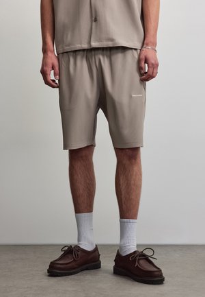Homme portant un short côtelé beige et une chemise assortie, des chaussettes blanches montant jusqu'à la cheville, et des chaussures à lacets marron foncé, debout sur un sol gris.