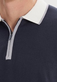 Granatowa koszulka polo z białą i szaro-rybkową kołnierzykiem oraz zamkiem błyskawicznym do ćwiartki. Materiał wydaje się być miękki i dziany o gładkiej fakturze.