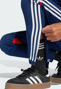 Marineblå träningsbyxor med vita ränder, röda detaljer och dragkedjefunktioner. Matchas med svarta Adidas-skor och svarta randiga strumpor.