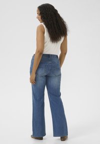 Vrouw met lang krullend haar, gekleed in een witte mouwloze top en blauwe flared jeans, staand en zijwaarts gekeerd op een effen achtergrond.