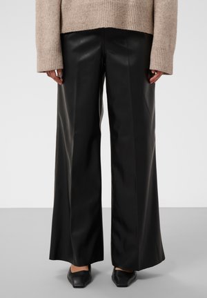 Personne portant un pantalon en cuir noir à jambes larges, un pull beige à manches longues et des chaussures noires pointues, debout devant un fond uni.