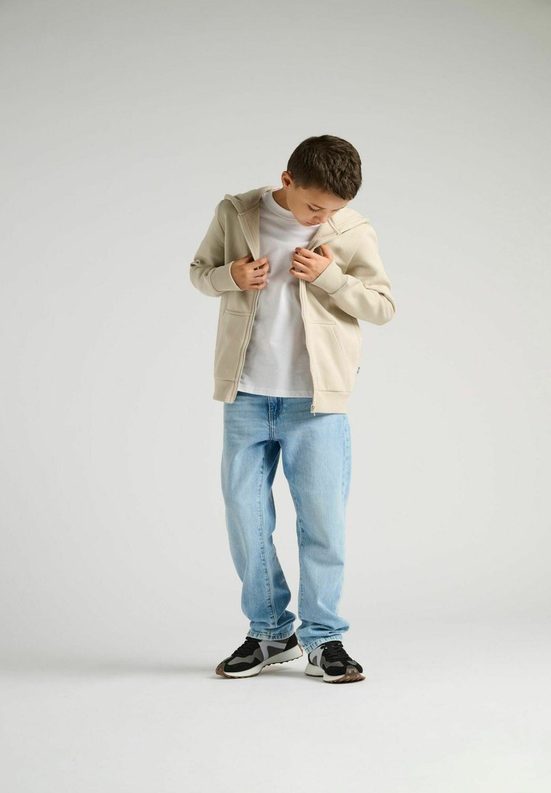 Felpa beige con zip, indossata con una t-shirt bianca sotto, abbinata a jeans azzurri chiari e sneakers nere. Design semplice, vestibilità casual.