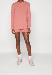 Person trägt ein passendes, staubrosa langärmeliges Hemd und Shorts mit weißen Socken und pinken Turnschuhen und steht vor einem einfarbigen Hintergrund.