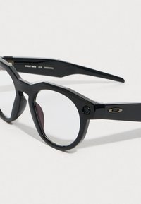 Schwarze rechteckige Sonnenbrille mit klaren Gläsern. Dicke Kunststofffassung mit eckigen Kanten und einer kleinen Kamera an der Seite. Silbernes Logo-Element.