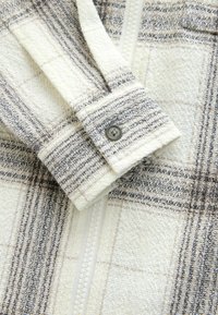 Koos nuhviga mansetiga kreklevalge ja halli ruudulise tekstuuriga jopevarrukas lähedalt vaadatuna, ees valge tõmblukk.