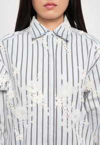 Chemise à rayures bleu et blanche avec un col, ornée de broderies en dentelle florale et de délicates perles sur le devant.