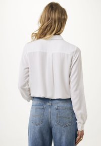 Blusa bianca con maniche lunghe, tessuto morbido e polsini bottonati, abbinata a jeans blu a vita alta con due tasche posteriori.
