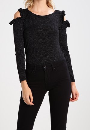 Femme portant un haut noir pailleté à manches longues avec découpes aux épaules et nœuds, associé à un pantalon noir ajusté.
