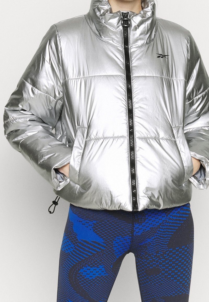 Person trägt eine glänzende silberne Steppjacke mit Händen in den Taschen, kombiniert mit blau-schwarz gemusterten Leggings.