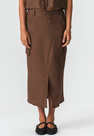 Žena v hnedej midi sukni s predným rozparkom a bočnými vreckami, v kombinácii s čiernymi topánkami Mary Jane, stojaca pred jednoduchým pozadím.