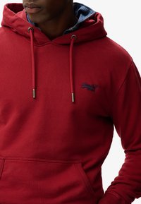 Felpa rossa realizzata in tessuto morbido con tasca a marsupio, cappuccio con coulisse e dettagli del colletto in denim blu. Presenta un logo di marca in blu.