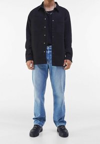 Zwarte button-up shirt met twee borstzakken, gedragen over een zwart T-shirt; vervaagde blauwe denimspijkerbroek; zwarte sneakers met een gestructureerd oppervlak.