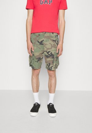 Personne portant un t-shirt rouge à manches courtes avec des inscriptions, un short cargo camouflage vert, des chaussettes blanches et des baskets noires sur un fond uni.