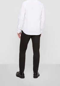 Camisa blanca de manga larga con un dobladillo curvado, combinada con pantalones negros ajustados y zapatos negros brillantes. Tejido suave y diseño minimalista.