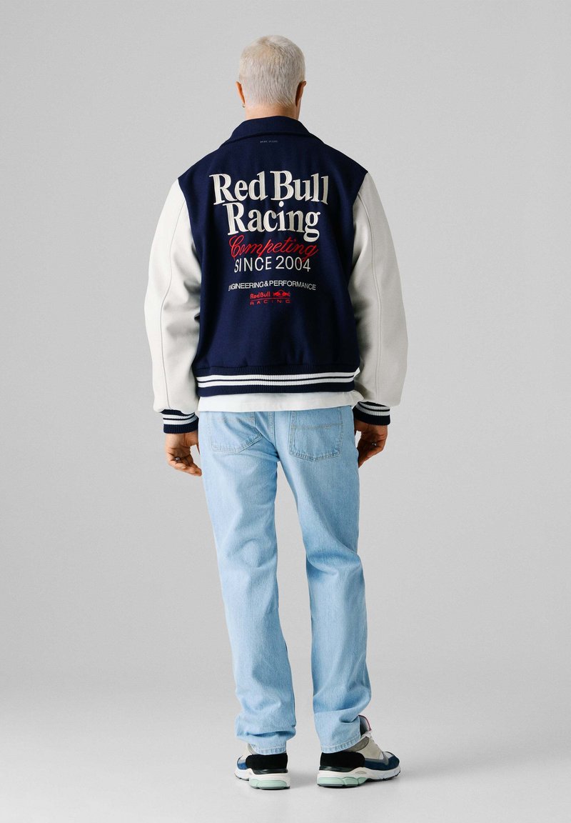Person med kort hvidt hår iført en marineblå og hvid Red Bull Racing-jakke, lyseblå jeans og sneakers, stående mod en grå baggrund.