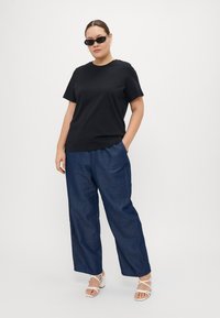 T-shirt nera in cotone a maniche corte, abbinata a pantaloni larghi in denim blu e sandali bianchi con cinturini. Modella che indossa occhiali da sole neri.