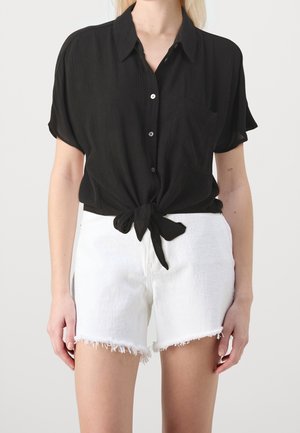 Femme portant une chemise noire à manches courtes boutonnée, nouée à la taille, et un short en jean blanc effiloché sur un fond uni.