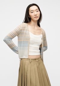 Νέα γυναίκα που φοράει μπεζ και μπλε πλεκτή ζακέτα, λευκό τοπάκι από κροσέ και ψηλόμεση πτυχωτή φούστα σε αποχρώσεις καφέ, στέκεται με τα χέρια πίσω από την πλάτη.