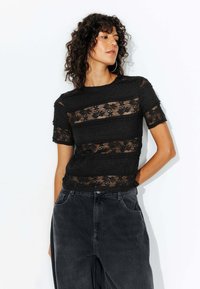 Haut en dentelle noire avec des bandes horizontales de tissu texturé, des manches courtes et un col ras du cou, associé à un pantalon en denim sombre.