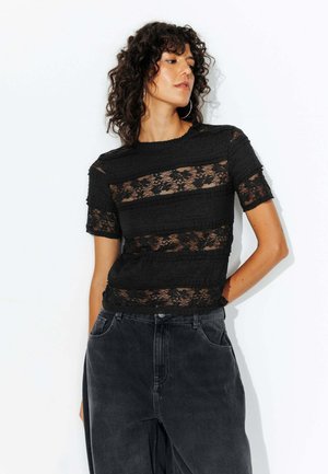 Haut en dentelle noire avec des bandes horizontales de tissu texturé, des manches courtes et un col ras du cou, associé à un pantalon en denim sombre.