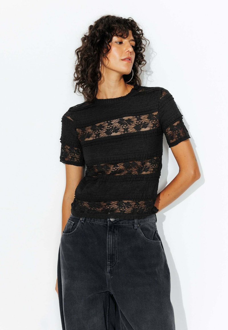 Haut en dentelle noire avec des bandes horizontales de tissu texturé, des manches courtes et un col ras du cou, associé à un pantalon en denim sombre.