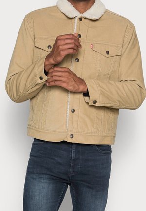 Homme ajustant le revers d'une veste en velours côtelé beige avec col sherpa blanc, portant un jean bleu foncé, sur un fond clair uni.