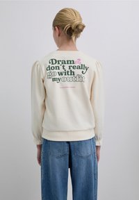 Hanorac crem cu mâneci bufante, cu un imprimeu mare de text grafic în verde și roz pe spate. Mâneci și tiv cu rebră.