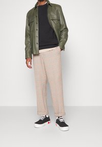 Giacca di pelle verde oliva sopra un maglione nero, pantaloni a quadri beige con dettagli arancioni e sneaker nere con suola bianca e dettagli rossi.
