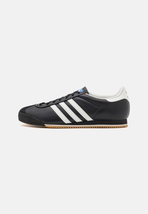 Sort lædersneaker med hvide striber, gummisål og sorte snørebånd, set fra siden på en ensfarvet lys baggrund.