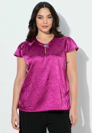 Kvinde med langt mørkt hår iført en skinnende magenta bluse med en dekorativ spænde ved halsudskæringen og sorte bukser, stående foran en hvid baggrund.