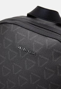 Black textured leather backpack featuring embossed logo and triangle patterns, with a smooth top handle and zip closure.